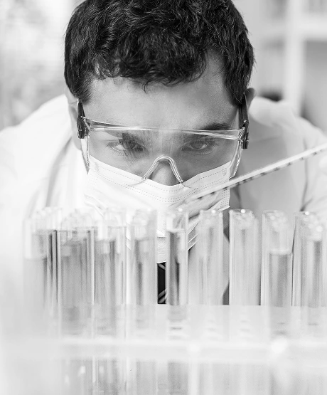 Scientist wearing protective goggles and a face mask closely examining a set of test tubes in a laboratory.